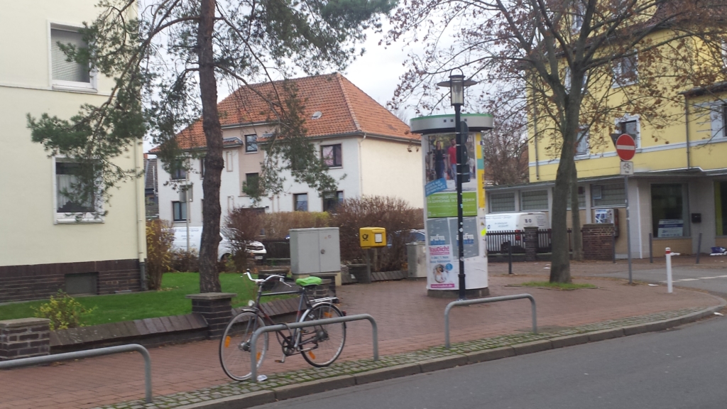 Ein Platz in Hildesheim. Auf dem Platz steht eine Litfaßsäule an einer Straße. Daneben steht ein Fahrrad. Im Hintergrund sind Wohnhäuser zu sehen.