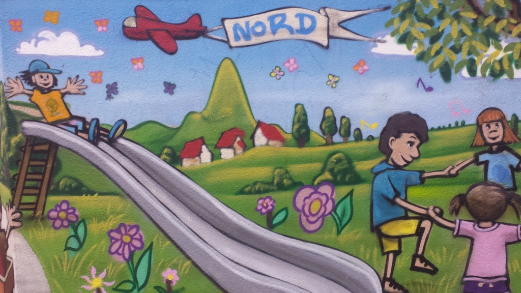 Eine Mauer, auf der ein Bild gemalt ist. Das Bild zeigt ein Kind auf einer Rutsche. Rechts im Bild sind weitere Kinder, die zusammen tanzen. Über den Kindern fliegt ein rotes Flugzeug. Das Flugzeug zieht ein Banner hinter sich her. Auf dem Banner steht "Nord".