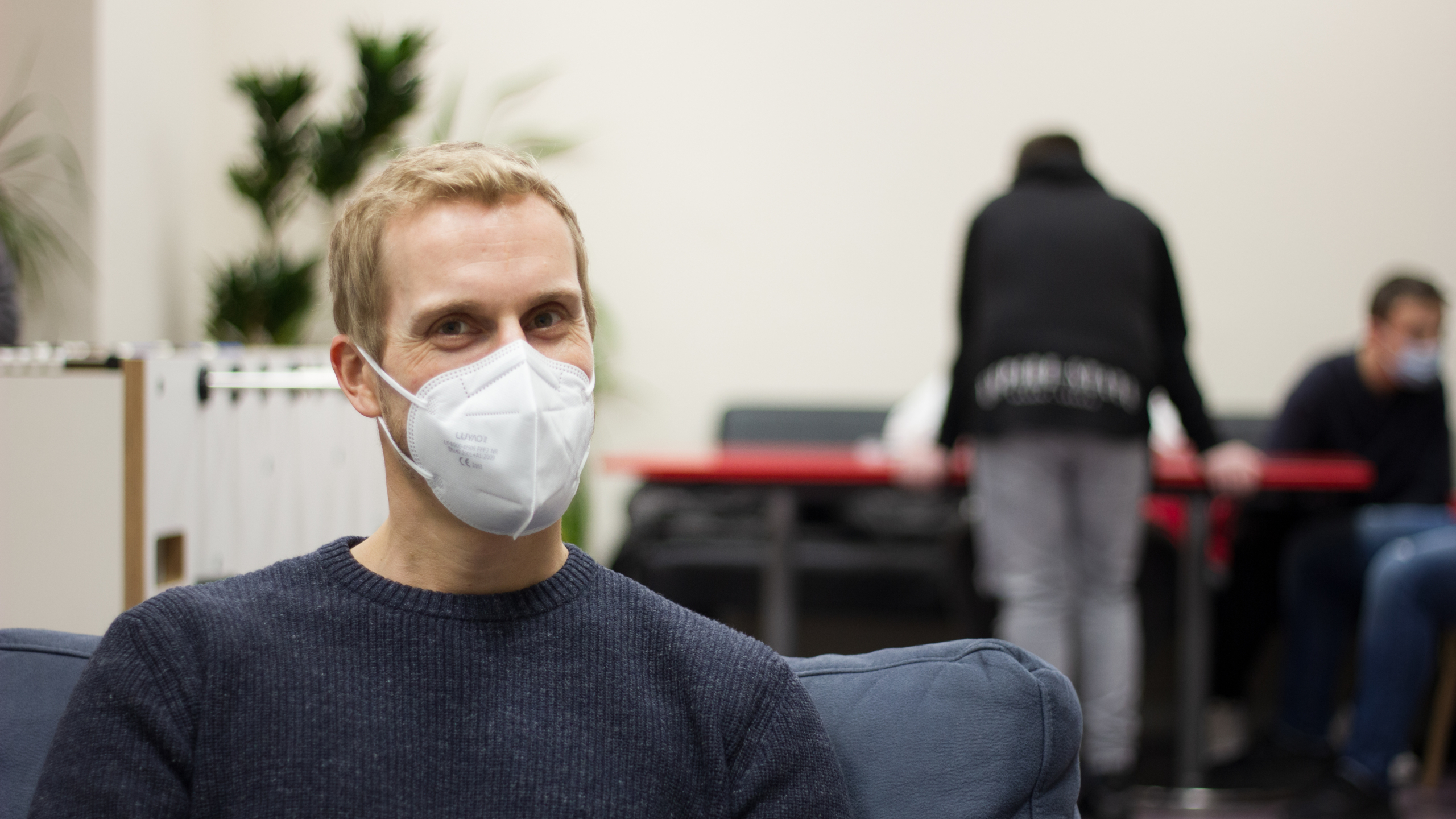 Ein Mann mit Corona-Maske lächelt in die Kamera. Im Hintergrund sind weitere Menschen verschwommen zu sehen.