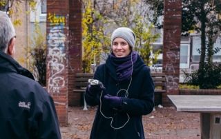 Eine Frau unterhält sich mit einem Mann. Sie hat ein Aufnahmegerät in der Hand. Im Hintergrund sieht man eine Tischtennisplatte.