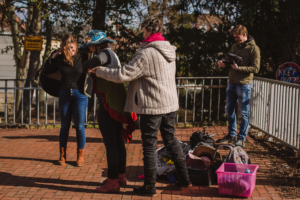 Eine Frau hilft einer anderen Frau dabei, ein Kostüm anzuziehen. Im Hintergrund steht ein Mann mit einer Kamera. Links steht eine weitere Frau und zieht eine Jacke an.