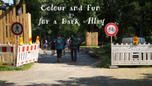 Menschen gehen einen Weg entlang. Links und rechts befindet sich eine Baustelle. Über dem Bild steht in weißer Schrift: "Colour and Fun for a Dark Alley".