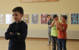 Kinder proben in der Schule ein Theaterstück. Ein Junge mit verschränkten Armen befindet sich links im Bild. Rechts stehen drei weitere Jungs und formen mit den Händen ein Kissen unter ihrem Kopf.