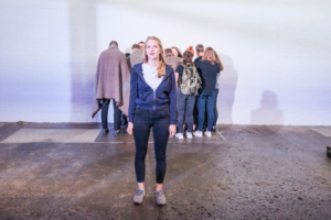 Eine junge Frau steht in der Mitte eines Raumes und blickt in die Kamera. Im Hintergrund befindet sich eine Gruppe von Jugendlichen. Sie stehen mit dem Rücken zu den Betrachtenden und haben die Köpfe gesenkt.