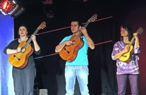 Drei Personen mit Gitarren stehen nebeneinander. Sie schauen starr nach vorne.