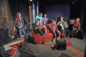 Viele Personen mit Musikinstrumenten sitzen in einem Raum.