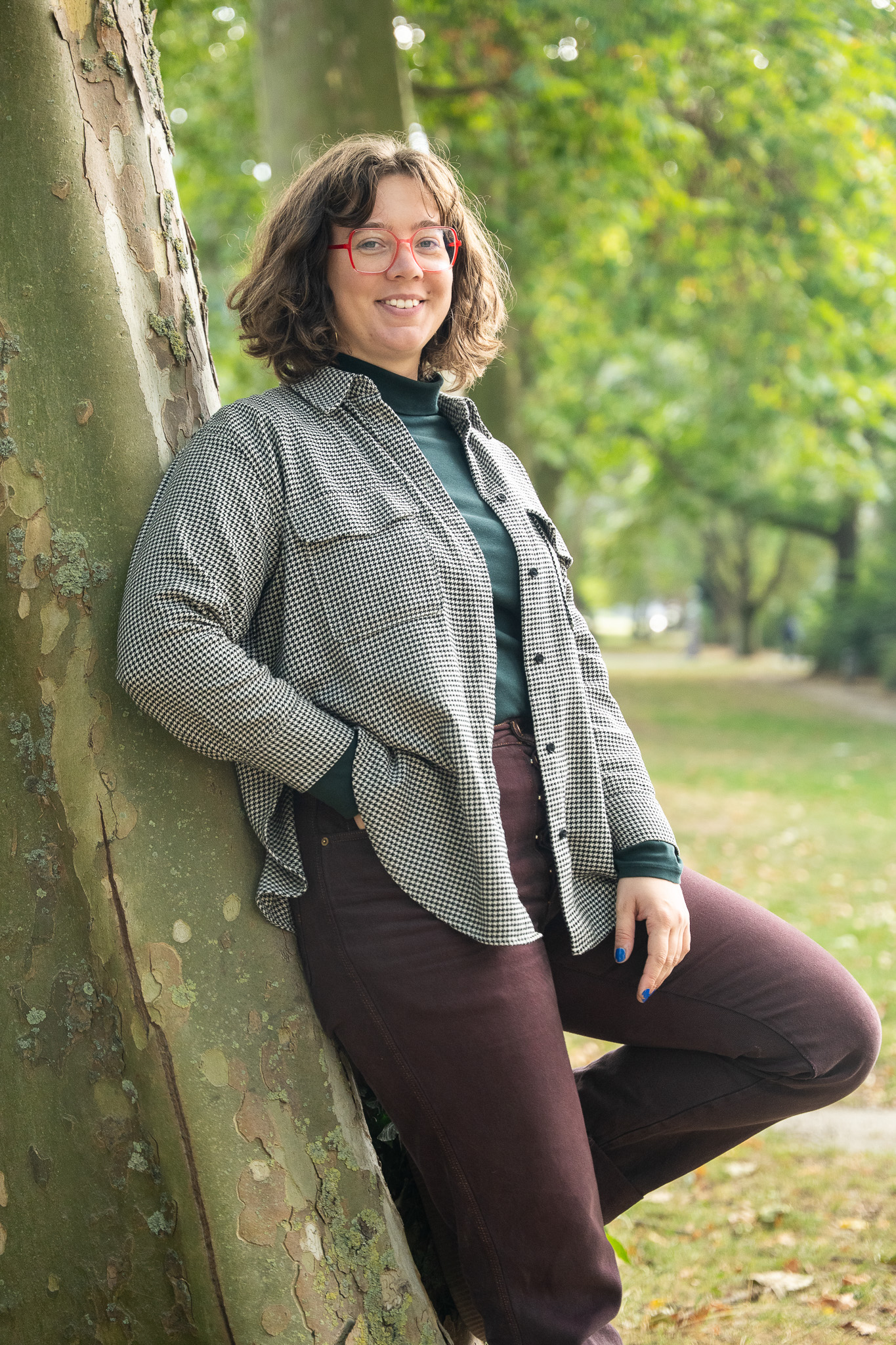 ine Frau lehnt am Baum und lacht in die Kamera. Sie trägt ein graues Hemd und eine braune Hose. Sie stützt sich mit einem Bein am Baum ab.