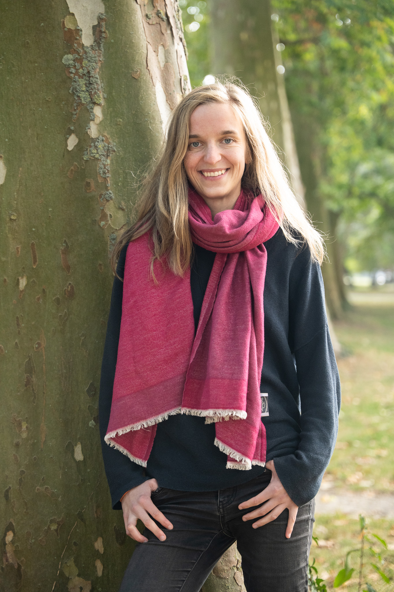 Eine Frau lehnt am Baum und lächelt in die Kamera. Sie trägt einen großen, rosa Schal.
