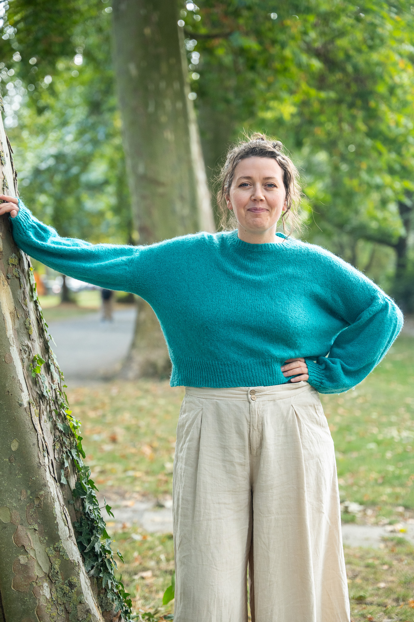 Eine Frau lehnt am Baum und lächelt in die Kamera. Sie stützt sich mit einem Arm am Baum ab. Sie trägt einen blauen Pullover und eine weiß-graue Hose.