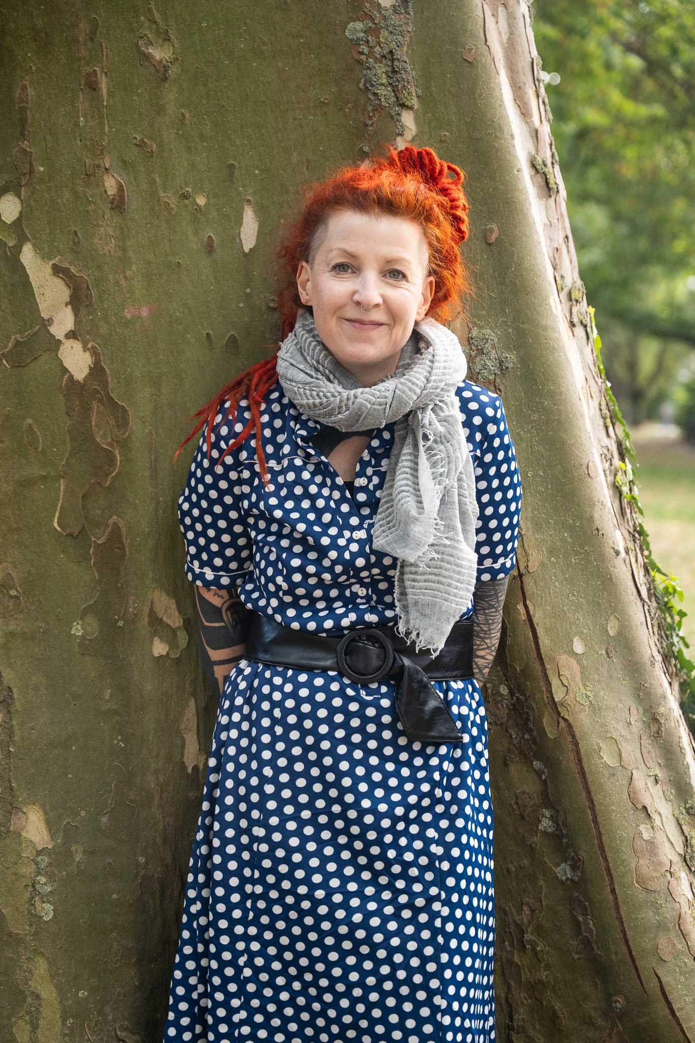 Eine Frau lehnt am Baum und lächelt in die Kamera. Sie trägt ein blaues Kleid mit weißen Punkten und einen weißen Schal.