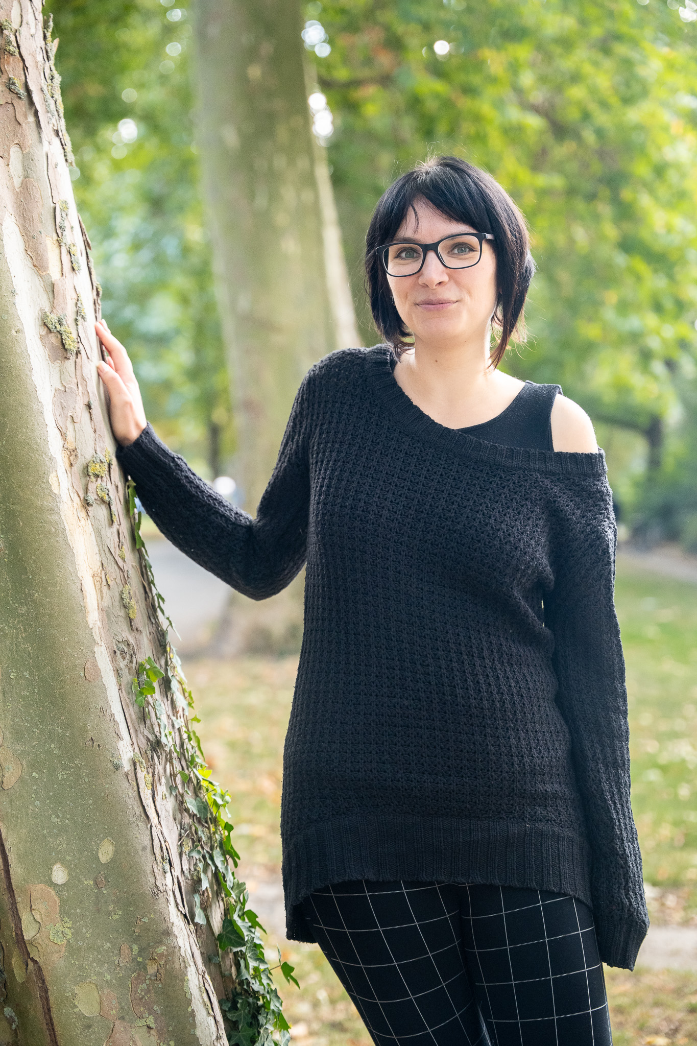 Eine Frau lehnt am Baum und lächelt in die Kamera. Sie trägt einen schwarzen Pullover.
