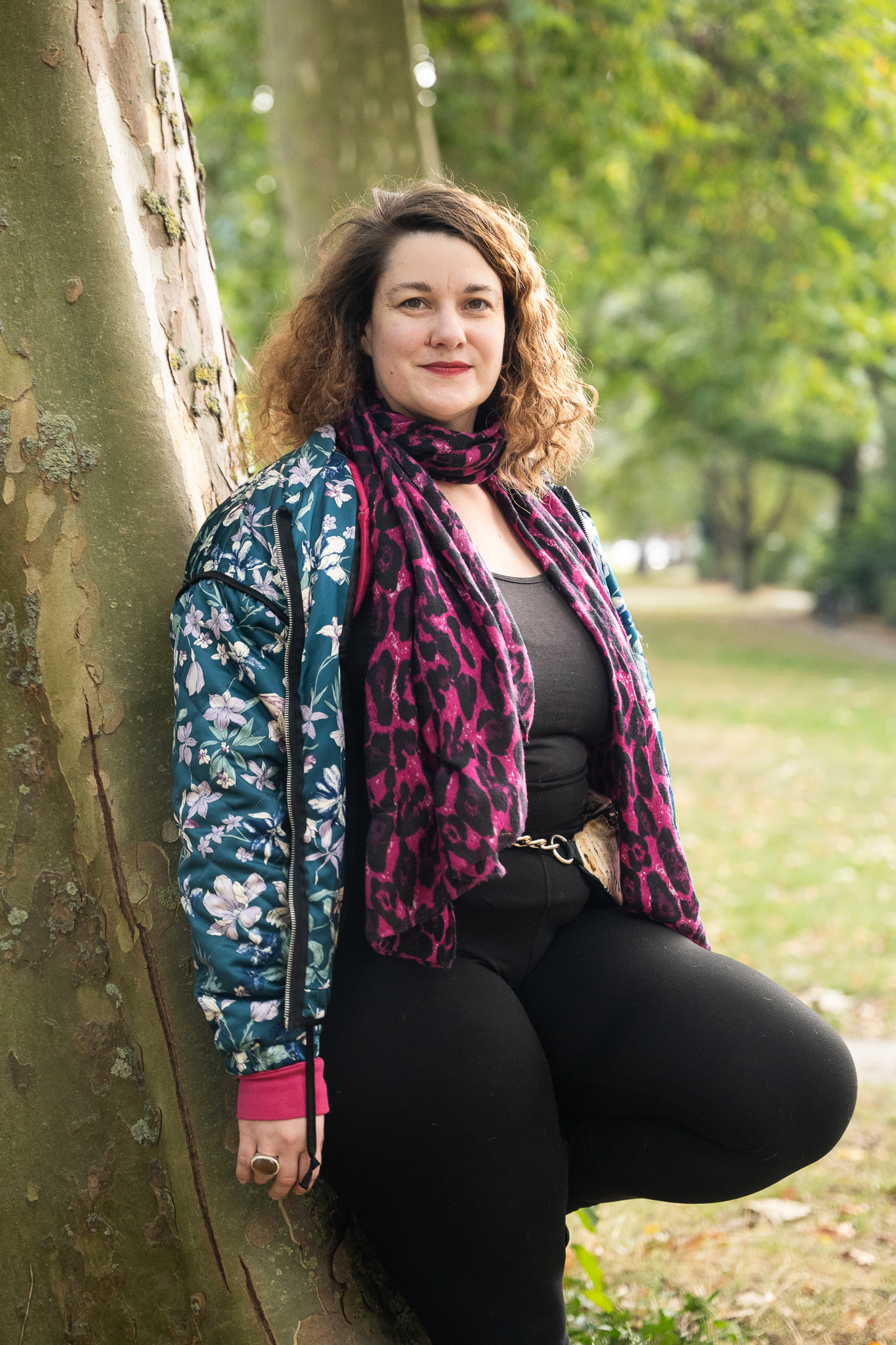 Eine Frau lehnt am Baum und lächelt in die Kamera. Sie trägt einen lila Schal mit Leopardenmuster und eine blaue Jacke mit einem Blumenmuster.