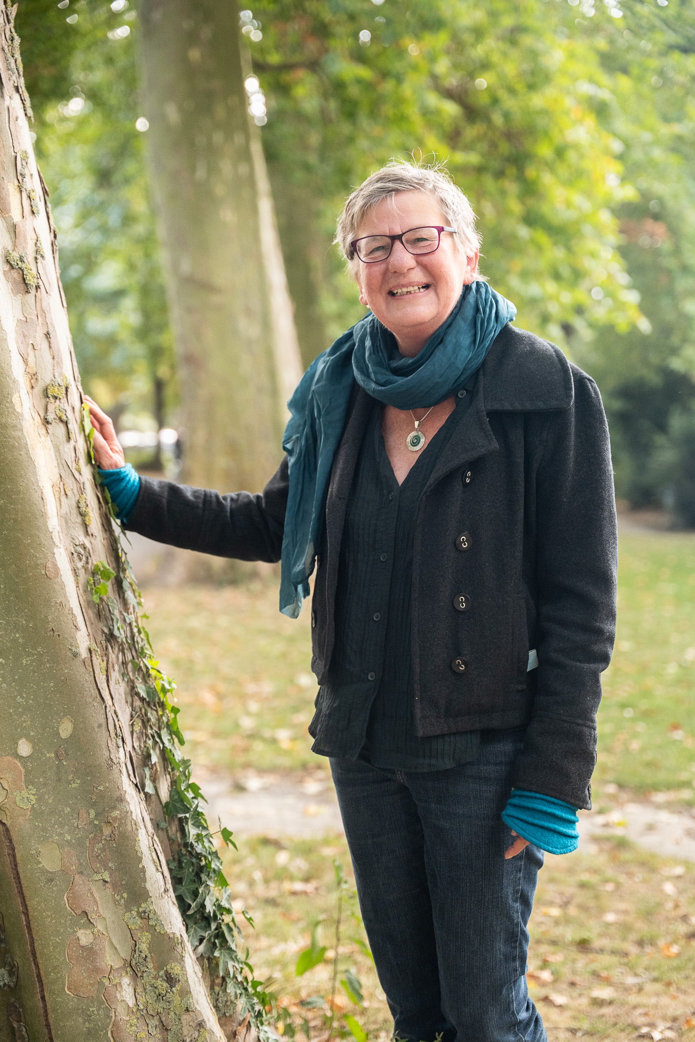 Eine Frau lehnt am Baum und lacht in die Kamera. Sie trägt einen blauen Schal und eine graue Jacke.