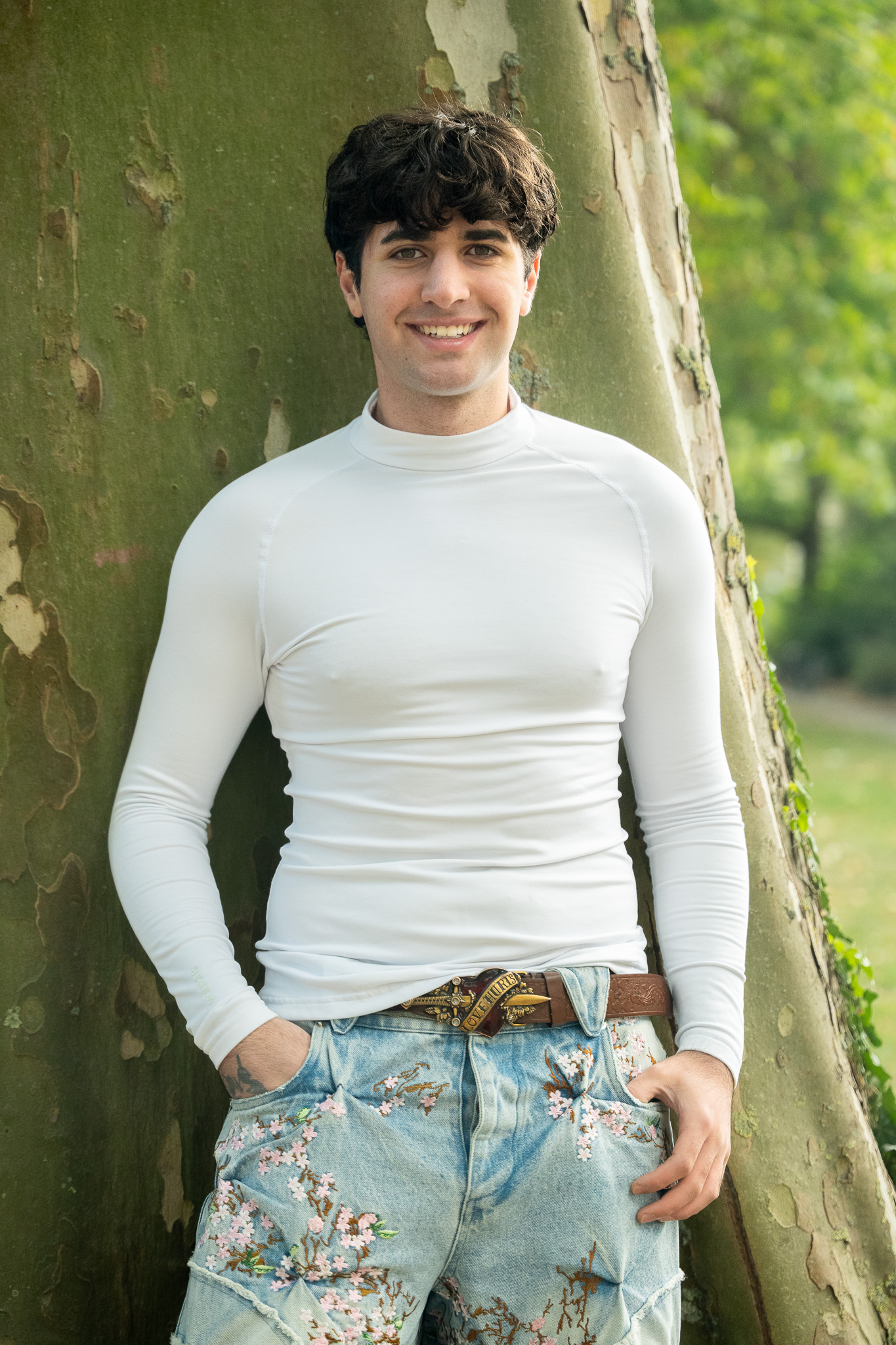Ein Mann lehnt an einem Baum und lächelt in die Kamera. Er trägt ein weißes Shirt und eine Jeanshose mit Blumenmuster.