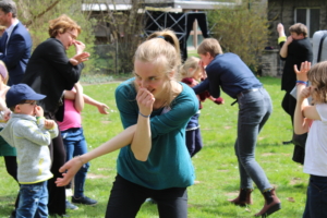 Eine junge Frau läuft über eine Wiese. Die Frau hat ihre Arme zu einem Elefantenrüssel geformt und lächelt. Im Hintergrund laufen Kinder und Erwachsene in gleicher Pose über die Wiese.