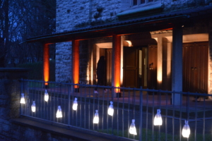 Die Türen der Martin-Luther-Kirche. Die Kirche ist blau-orange beleuchtet. Im Vordergrund befindet sich ein Zaun. Daran hängen mehrere weiße Beutel, die leuchten.