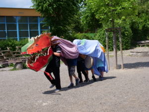Kinder tragen einen gebastelten Drachen. Der rote Kopf des Drachen besteht aus Pappe, der Körper besteht aus bunten Bettlaken.