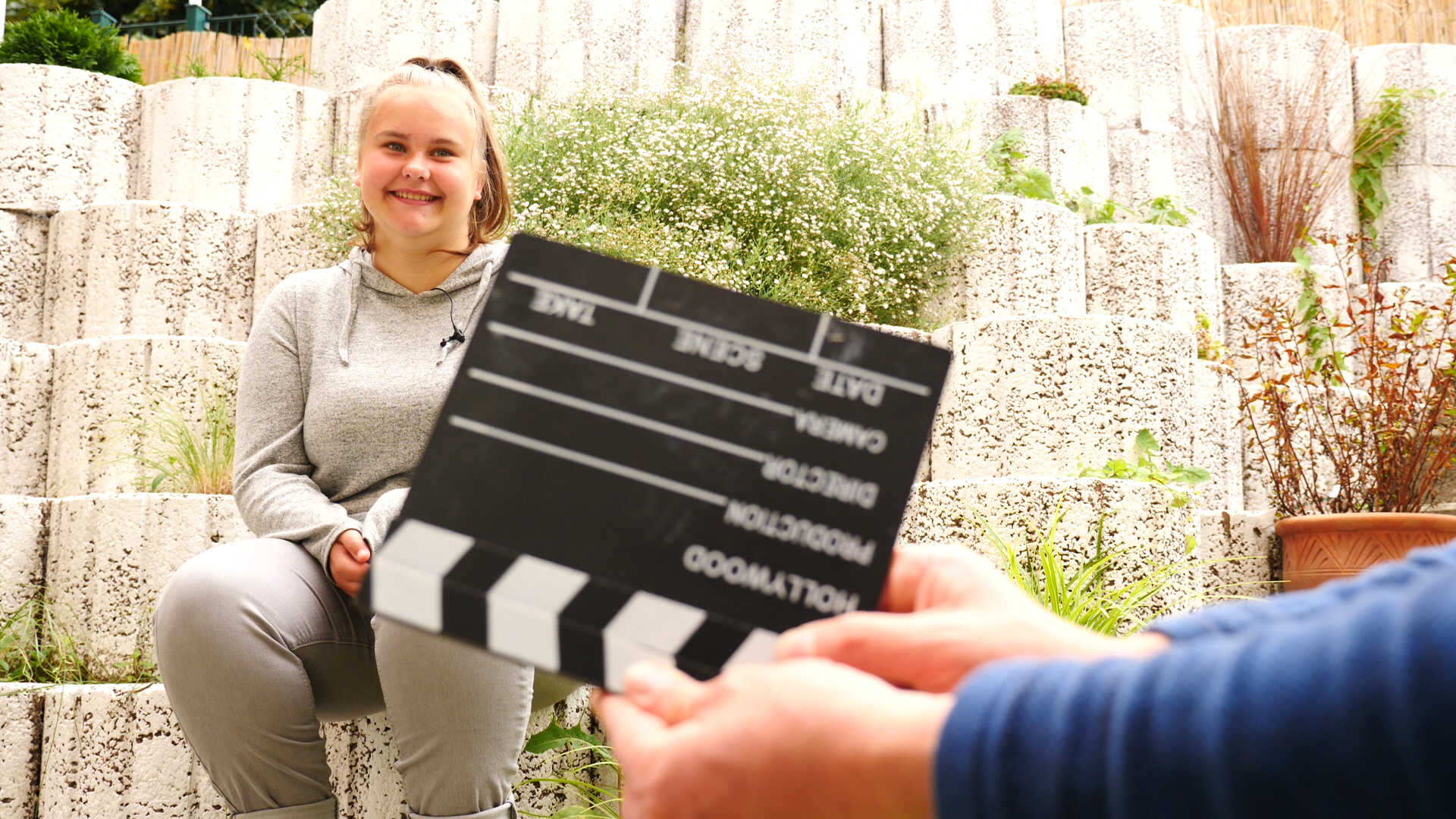 Eine junge Frau sitzt auf einem Stein und lächelt. Vor ihr hält eine Hand eine Filmklappe in die Kamera.