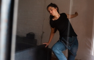 Eine Frau tanzt. Neben ihr steht ein schwarzes Ledersofa. Im Hintergrund befindet sich eine weiße Mauer.