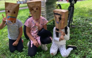 Kinder mit Papiermasken auf dem Kopf sitzen auf einer Wiese.