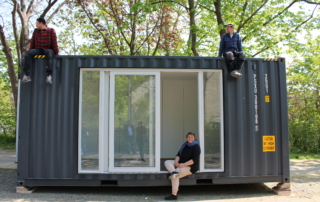 Ein Schiffscontainer mit verglaster Außenfront steht auf einer Straße. Auf dem Schiffscontainer sitzen zwei Männer und schauen nach links. Im offenen Fenster des Schiffscontainers sitzt eine Frau und lacht in die Kamera.