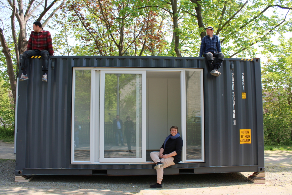 Ein Schiffscontainer mit verglaster Außenfront steht auf einer Straße. Auf dem Schiffscontainer sitzen zwei Männer und schauen nach links. Im offenen Fenster des Schiffscontainers sitzt eine Frau und lacht in die Kamera.