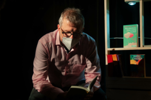Ein Mann sitzt vor einem Bücherregal. Er liest ein Buch. Der Hintergrund ist dunkel.