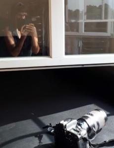 Auf einem schwarzen Ledersessel liegt eine Kamera. Darüber befindet sich ein offenes Fenster. In der Fensterscheibe spiegelt sich eine junge Frau mit einem Handy.