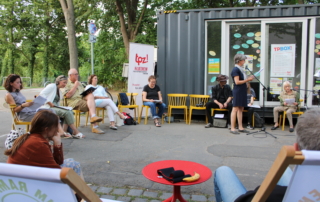 Menschen sitzen im Kreis vor einem Schiffscontainer. Eine Person liest etwas vor.