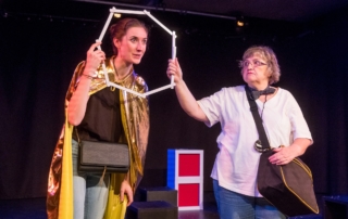 Eine Frau schaut durch einen Zollstock, den sie zu einem Kreis gebogen hat. Sie trägt einen goldenen Umhang. Neben ihr steht eine ältere Frau in einem weißen T-Shirt. Sie hält den Zollstock mit einer Hand fest.