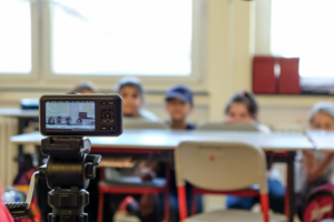 Kinder verstecken sich hinter einer Schulbank. Im Vordergrund steht eine Kamera und filmt die Kinder.