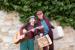 Eine Frau und ein Mann mit Gitarre und Koffern in der Hand schauen fröhlich in die Kamera.