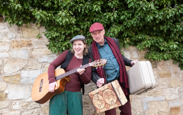 Eine Frau und ein Mann mit Gitarre und Koffern in der Hand schauen fröhlich in die Kamera.