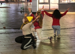 Drei Personen tanzen zusammen auf einem Bahnhofsvorplatz.