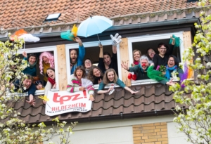 Viele verkleidete Menschen winken aus einem Fenster.