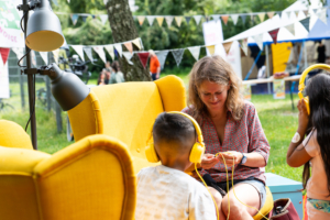 Eine Frau und zwei Kinder sitzen auf gelben Sesseln in einem Park. Die Kinder tragen Kopfhörer, sie sind nur von hinten zu sehen. Die Frau schließt das Kabel der Kopfhörer an einem Abspielgerät an.