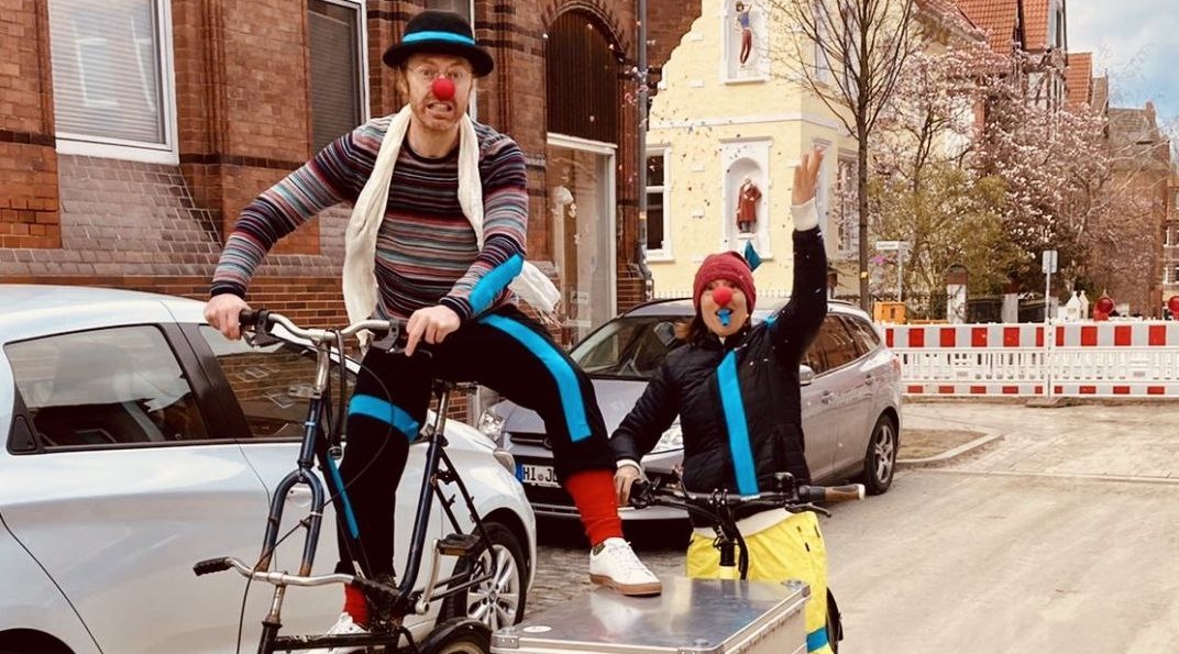 Zwei als Clowns verkleidete Theaterpädagog*innen fahren auf Fahrrädern durch Hildesheim.