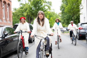 Vier Frauen, mit weißen Bademänteln bekleidet, fahren mit einem Fahrrad in Richtung der Kamera.