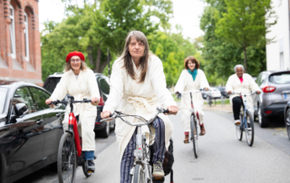 Vier Frauen, mit weißen Bademänteln bekleidet, fahren mit einem Fahrrad in Richtung der Kamera.