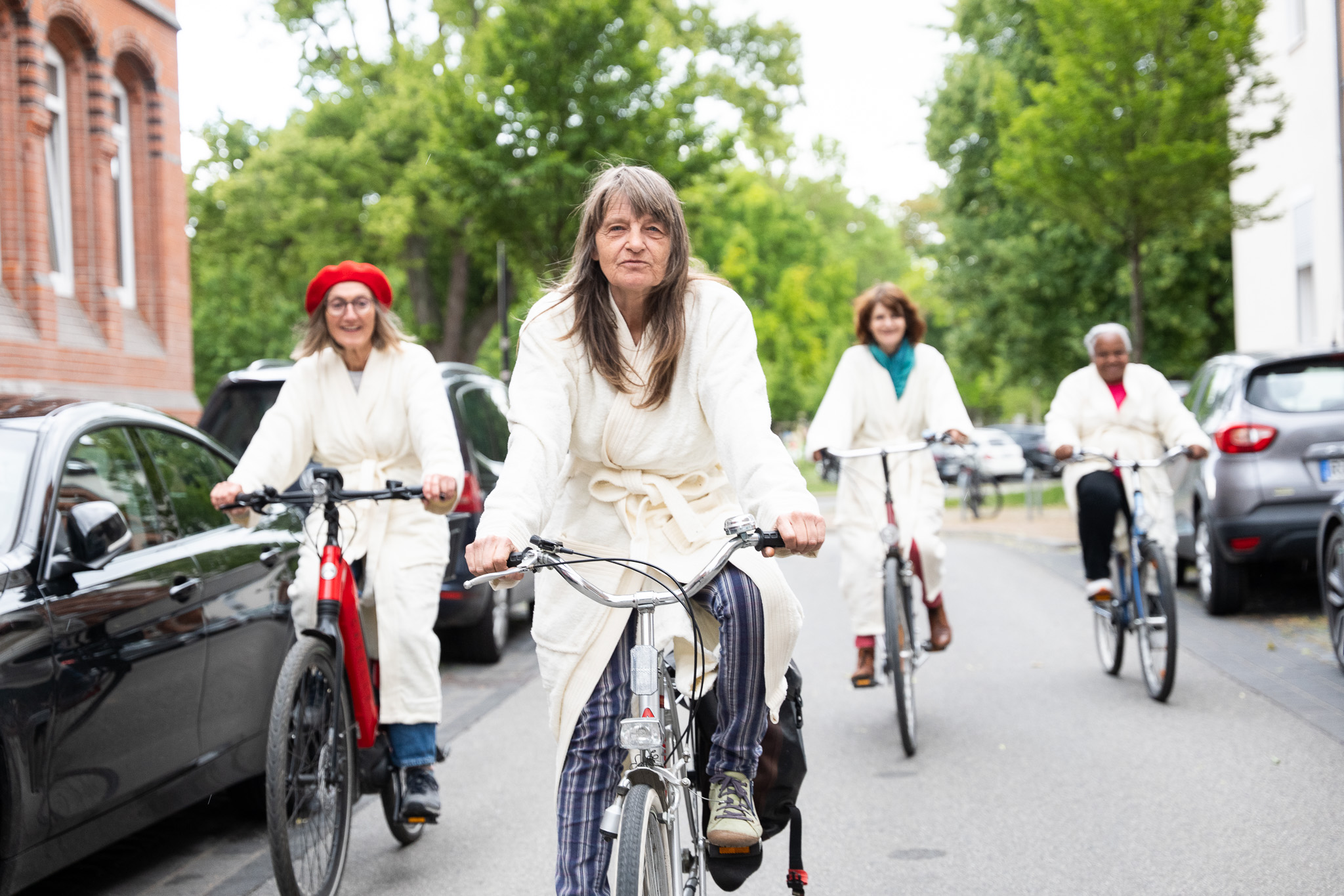 Vier Frauen, mit weißen Bademänteln bekleidet, fahren mit einem Fahrrad in Richtung der Kamera.