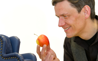 Ein Mann schaut lächelnd auf einen Apfel in seiner Hand. Neben ihm befindet sich ein kleiner, blauer Sessel.