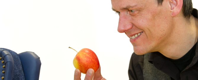 Ein Mann schaut lächelnd auf einen Apfel in seiner Hand. Neben ihm befindet sich ein kleiner, blauer Sessel.