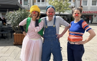 Drei als Clowns verkleidete Theaterpädagog*innen lachen in die Kamera.