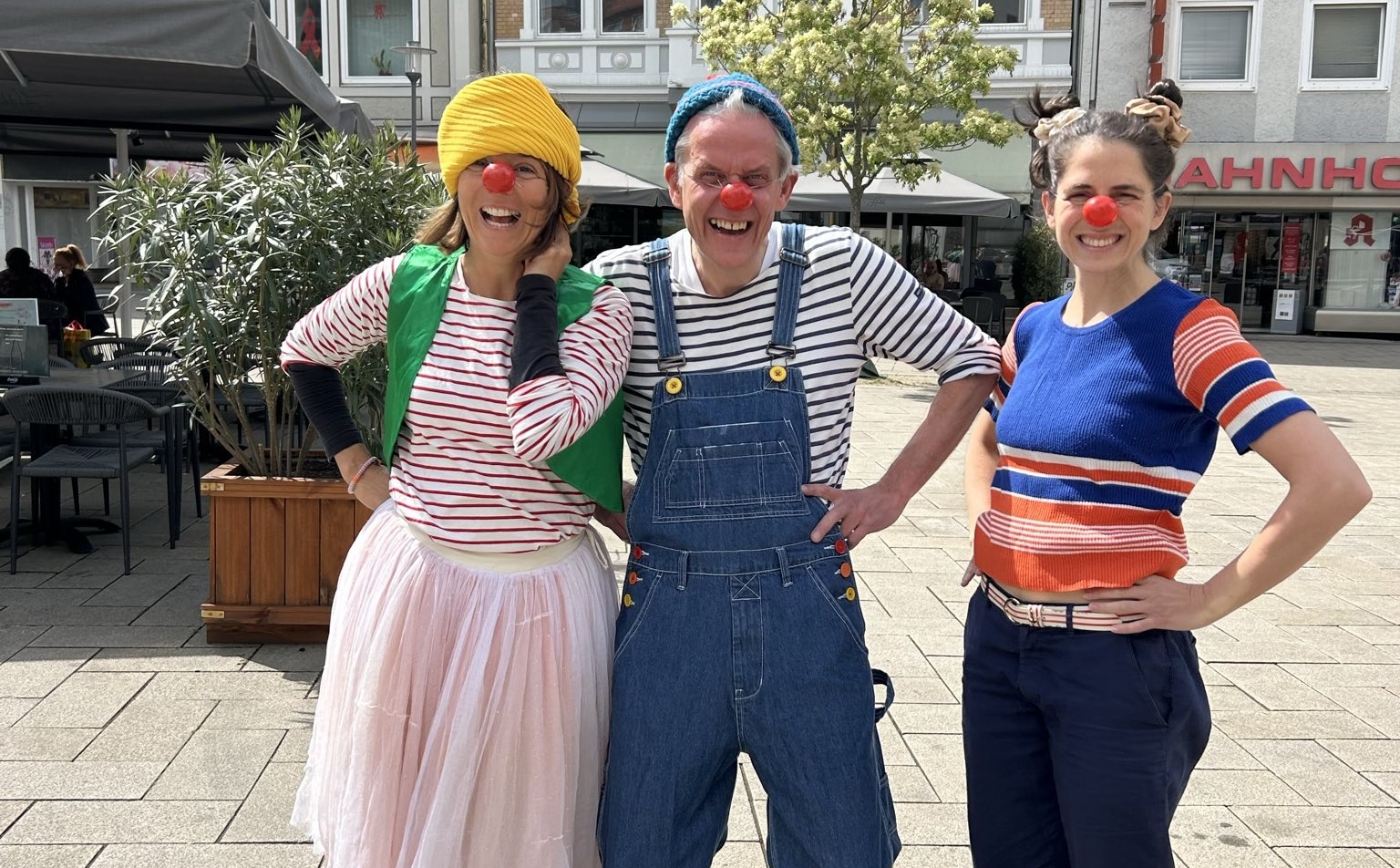 Drei als Clowns verkleidete Theaterpädagog*innen lachen in die Kamera.