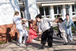 Zwei erwachsene Frauen und mehrere Kinder tanzen gemeinsam auf einem öffentlichen Platz im Kreis.