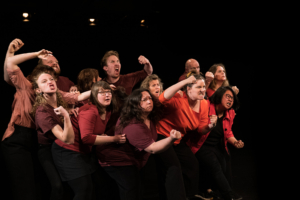 Viele Menschen stehen gemeinsam auf einer Bühne, recken die Fäuste in die Luft und blicken wütend in die gleiche Richtung.