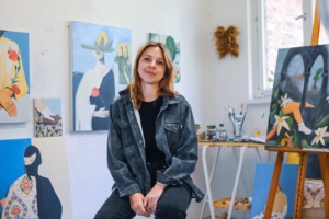 Eine junge Frau sitzt in einem Atelier, hinter ihr befinden sich viele gemalte Bilder.