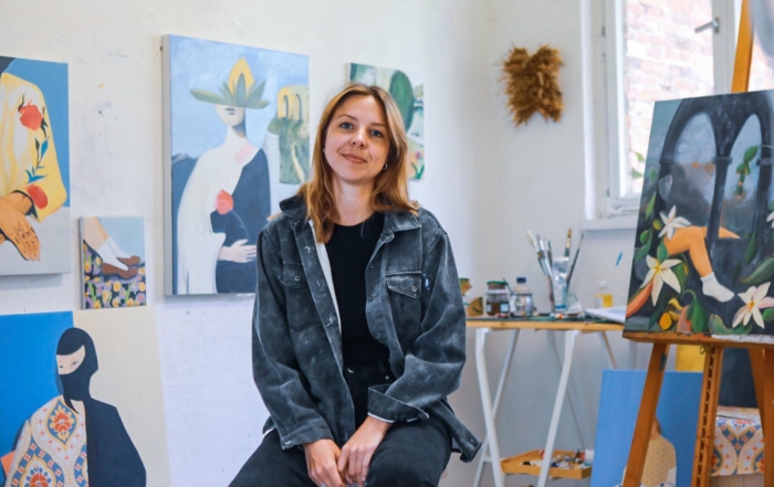 Eine junge Frau sitzt in einem Atelier, hinter ihr befinden sich viele gemalte Bilder.