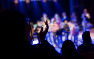 Kinder stehen im Pulk auf einer erleuchteten Bühne. Das Publikum applaudiert. Die Menschen sind nur als Schemen zu erkennen.