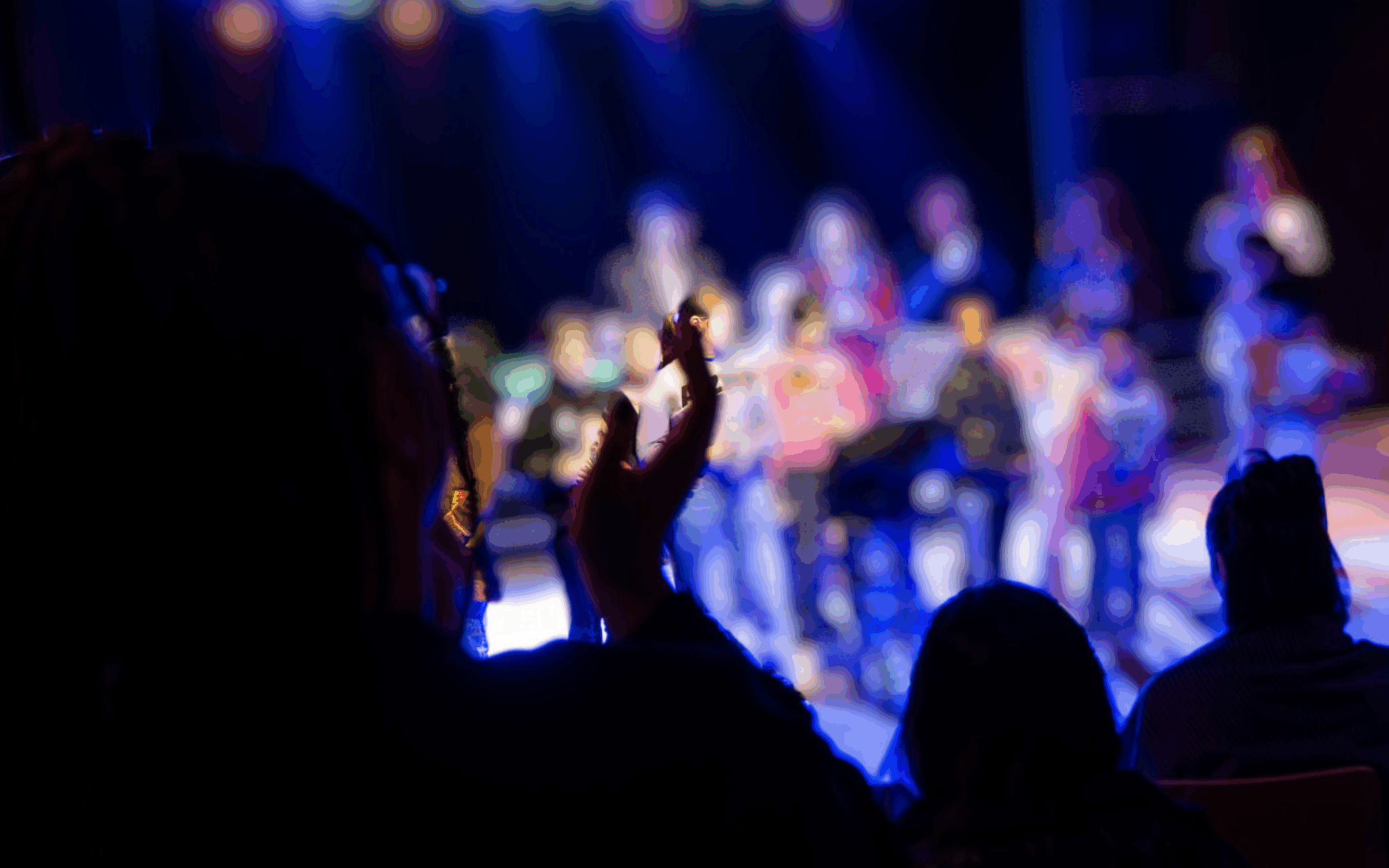 Kinder stehen im Pulk auf einer erleuchteten Bühne. Das Publikum applaudiert. Die Menschen sind nur als Schemen zu erkennen.